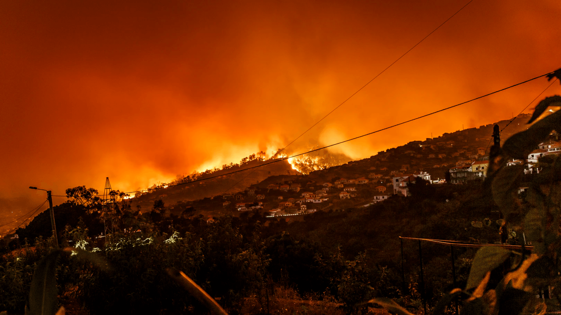 Scene of a wildfire on a hill at night. Houses are visible on the hillside. The sky is filled with an orange glow from the flames and smoke.